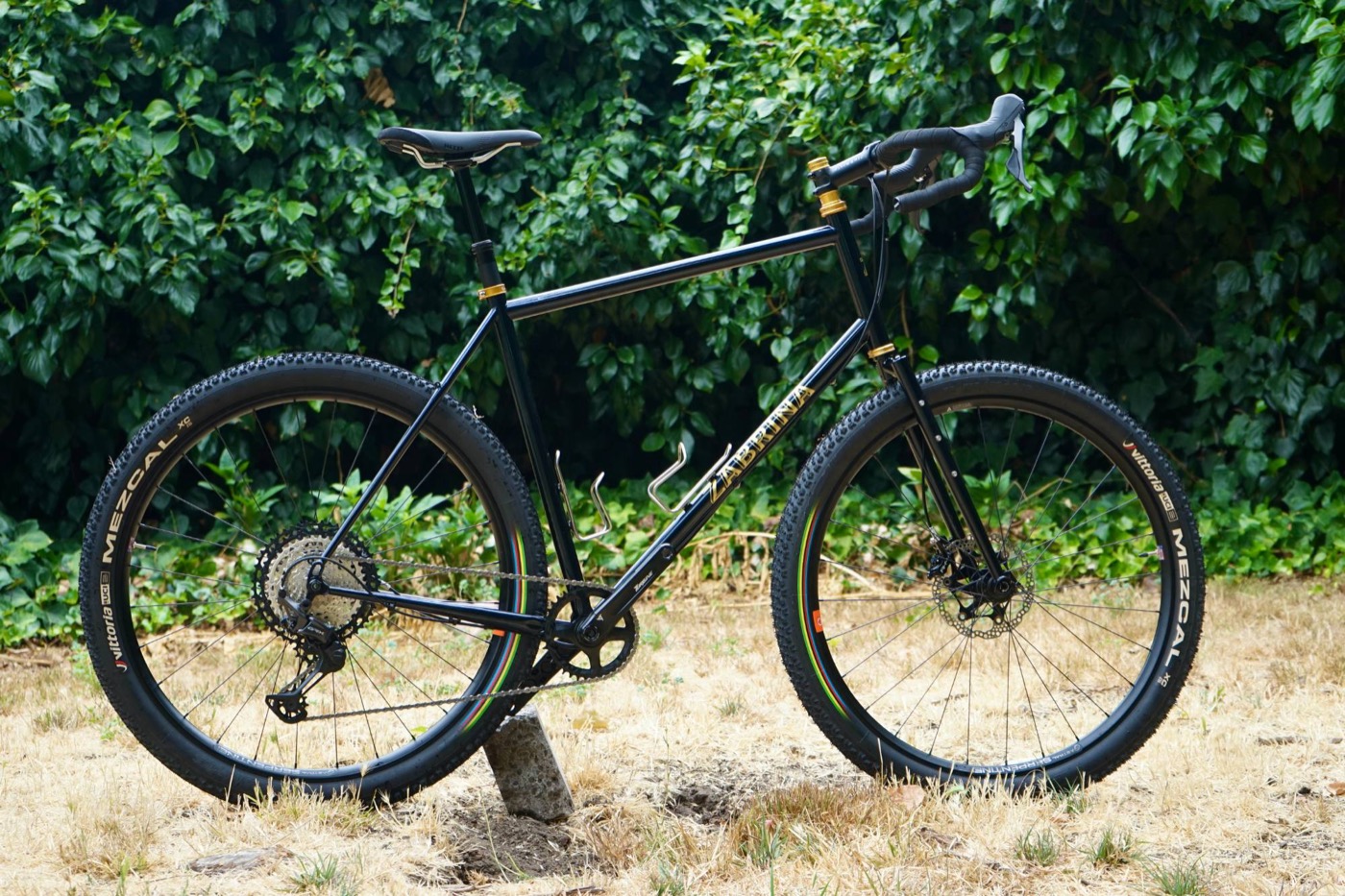 Midnight blue Zabrina drop-bar gravel bike with gold headset and knobby tires on dry grass