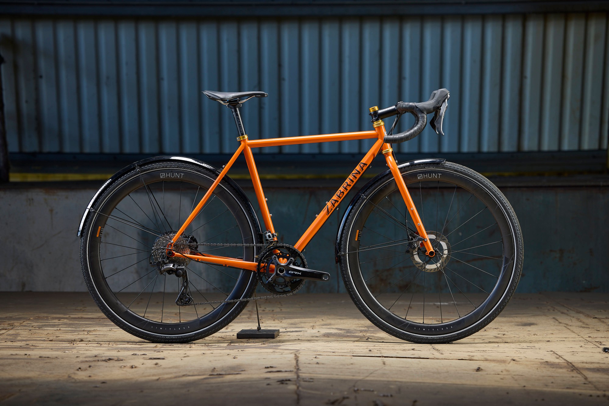Orange Zabrina road bike with black fenders and gold headset on a kickstand in front of a corrugated metal wall