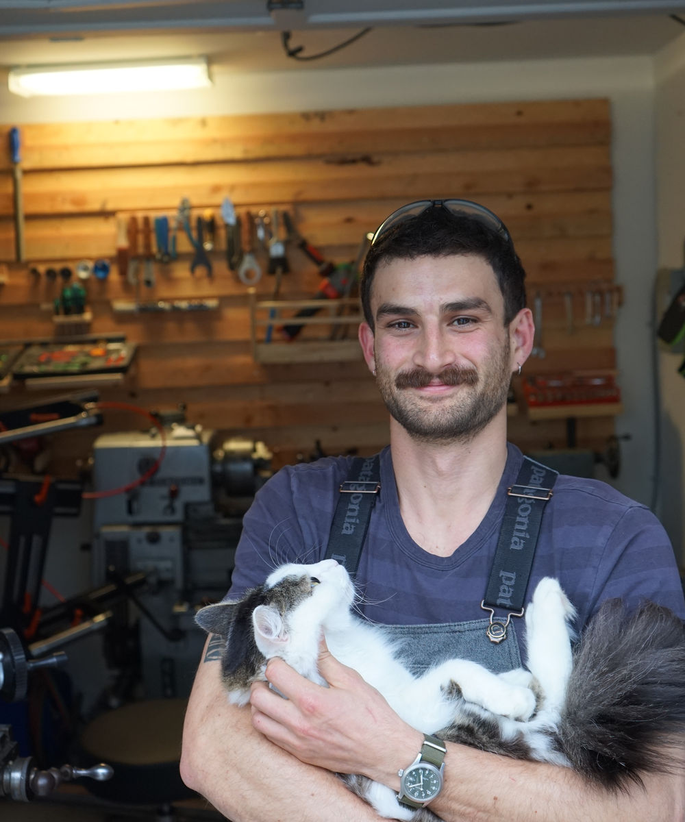 The builder in overalls holding a fluffy black-and-white cat in the Oakland shop