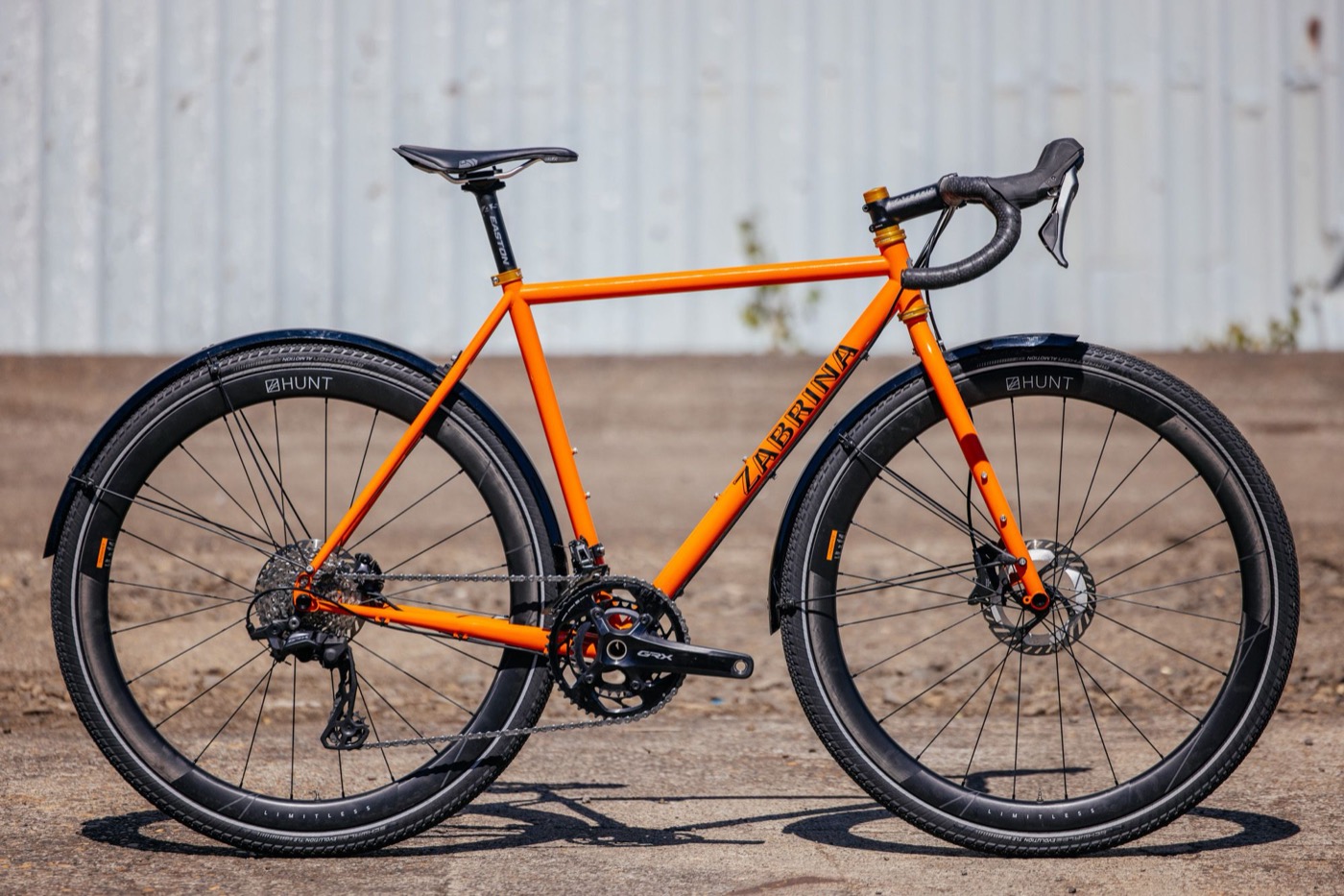 Orange Zabrina road bike with black fenders and gold headset in front of a corrugated wall