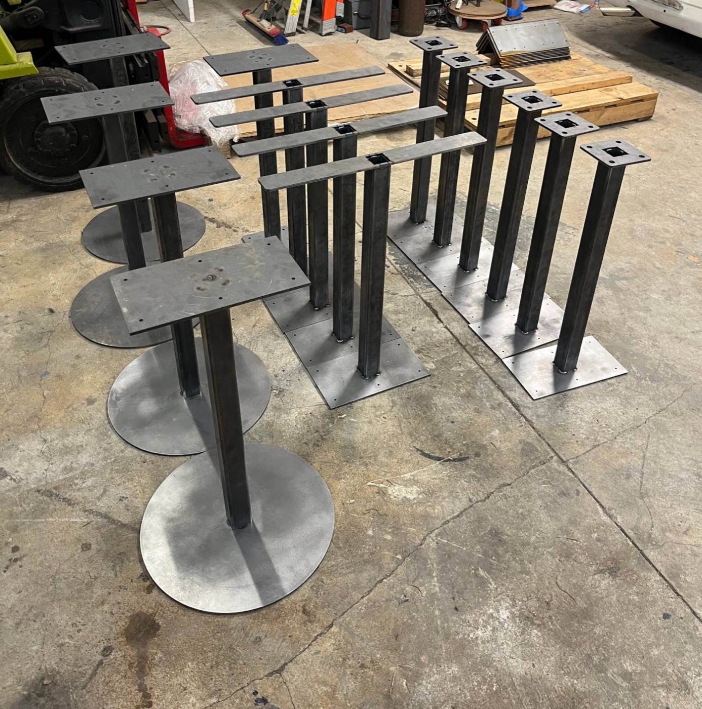 A dozen raw steel pedestal table bases with round and square feet on the shop floor
